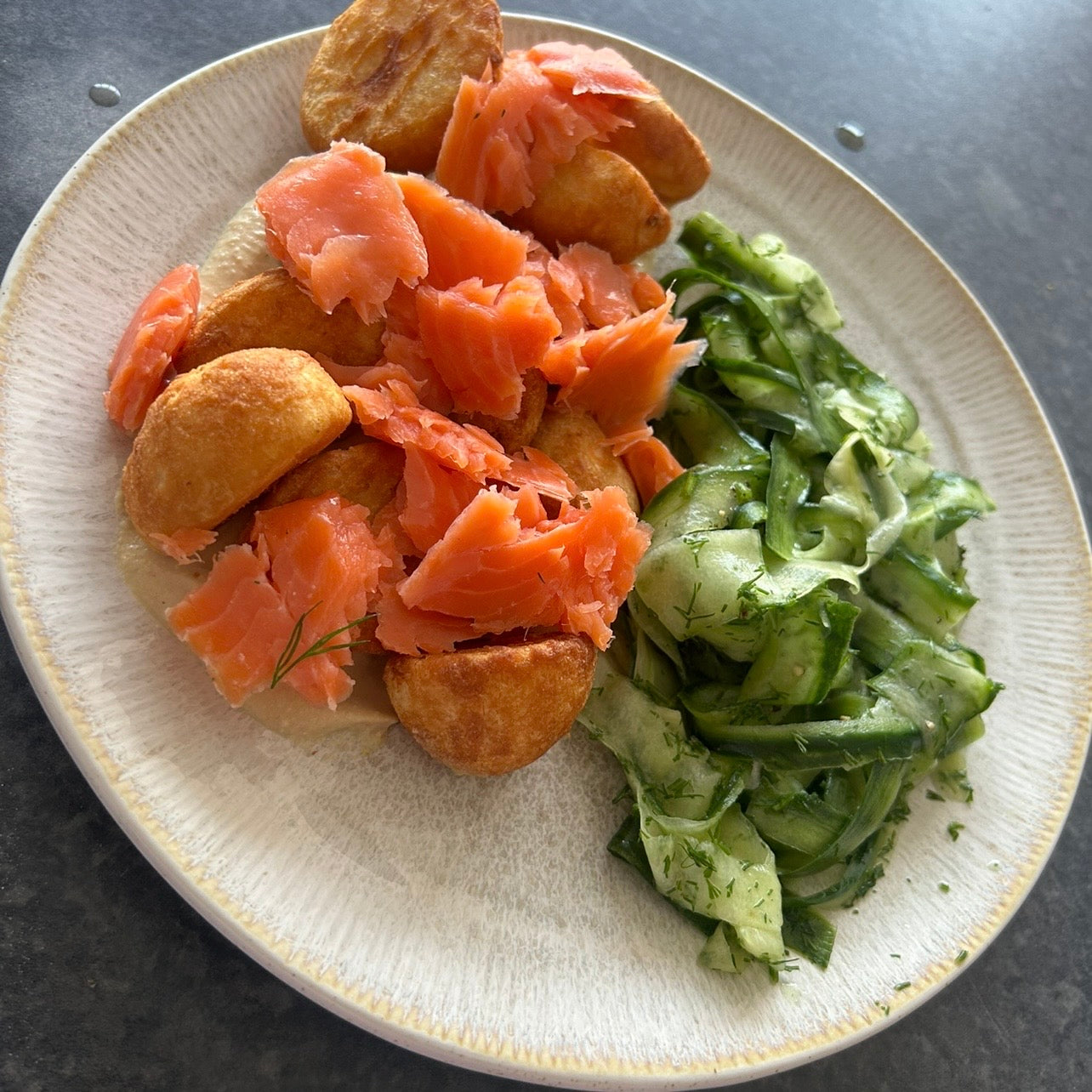 Bowl with crispy roasted potatoes, flaky smoked trout, creamy hummus and cucumber dill salad finished with lemon and olive oil.